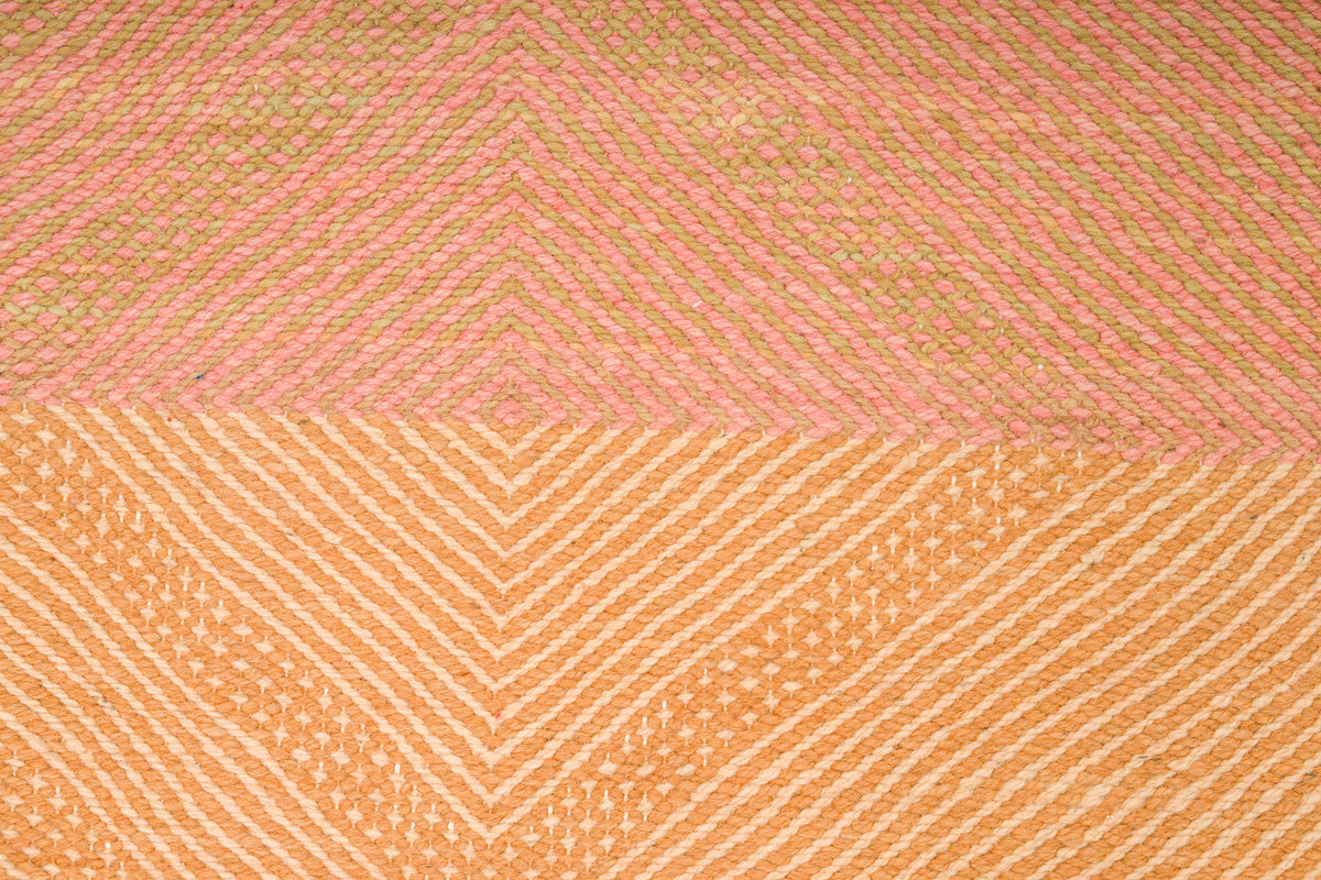 A close-up of a woven Zanafi rug with a geometric chevron pattern, split horizontally into two sections: a top section in dusty pink and a bottom section in warm peach-orange, both with fine striped texture.