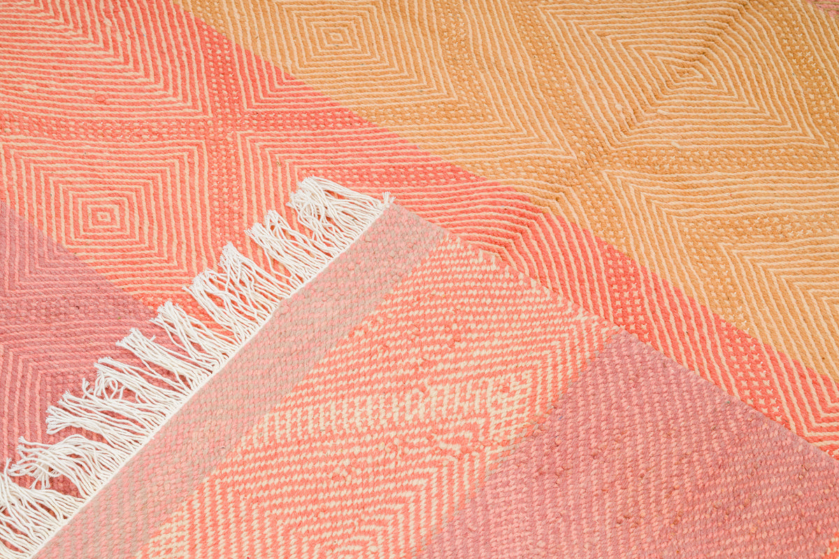A close-up of a Moroccan Zanafi Rug with geometric diamond patterns in coral pink, peach, and warm beige, finished with white fringe along one corner.