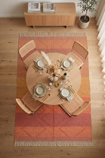 A top-down view of a round wooden dining table set with four woven cane chairs on a striped orange, rust, and beige Zanafi area rug with fringe, in a bright room with a light wood floor and a small wooden cabinet in the background.