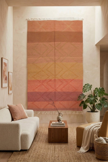 A large Zanafi woven rug  used as wall hanging or tapestry with horizontal terracotta, peach, orange, and muted pink stripes hangs on a beige wall in a styled living room with a sofa, coffee table, plant, and rug.