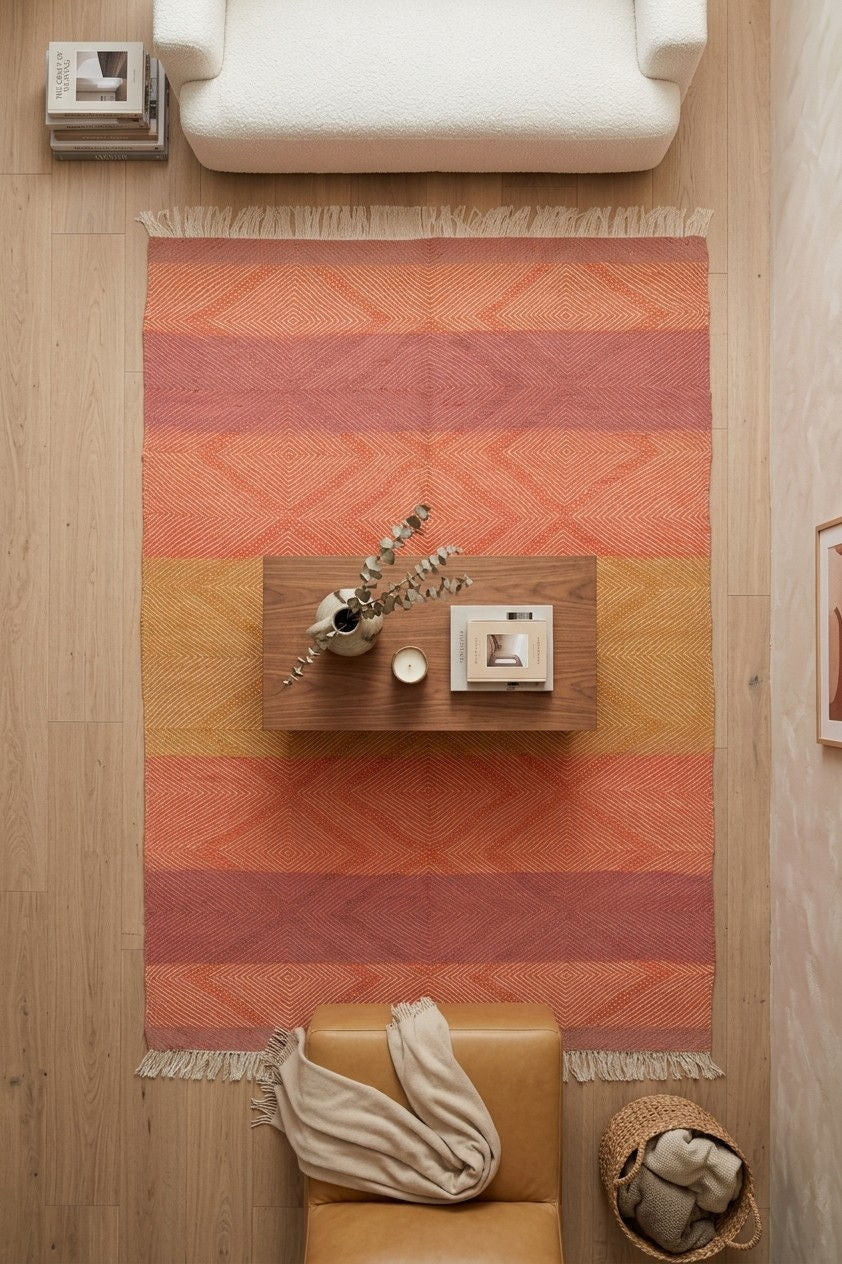A rectangular area rug with horizontal stripes in terracotta, orange, and mustard yellow, featuring a geometric diamond pattern and fringe on the ends. The rug is styled in a modern living room setting with a light wood floor, a white armchair at the top, a tan leather chair at the bottom, and a small floating wooden shelf in the center.