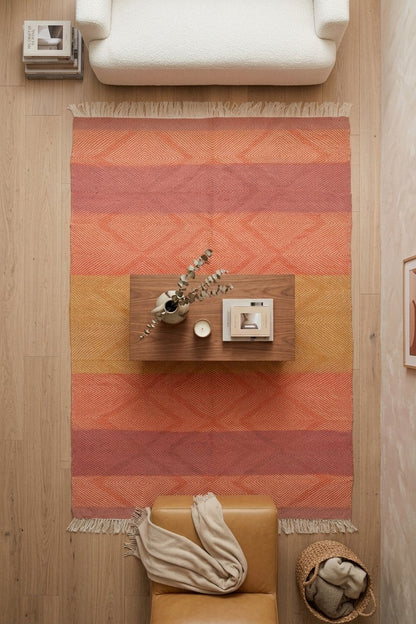 A rectangular area rug with horizontal stripes in terracotta, orange, and mustard yellow, featuring a geometric diamond pattern and fringe on the ends. The rug is styled in a modern living room setting with a light wood floor, a white armchair at the top, a tan leather chair at the bottom, and a small floating wooden shelf in the center.