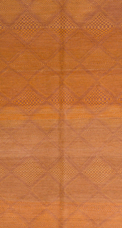 A close-up of a wool Moroccan rug in warm orange-brown tones, showing a repeating diamond pattern and subtle central seam.
