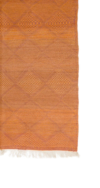 An orange woven textile with a geometric diamond pattern and white fringe along one edge, shown against a plain white background.