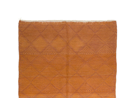 A square woven Zanafi rug in a warm rust-orange color, featuring a geometric diamond pattern and frayed edges, shown flat against a white background.