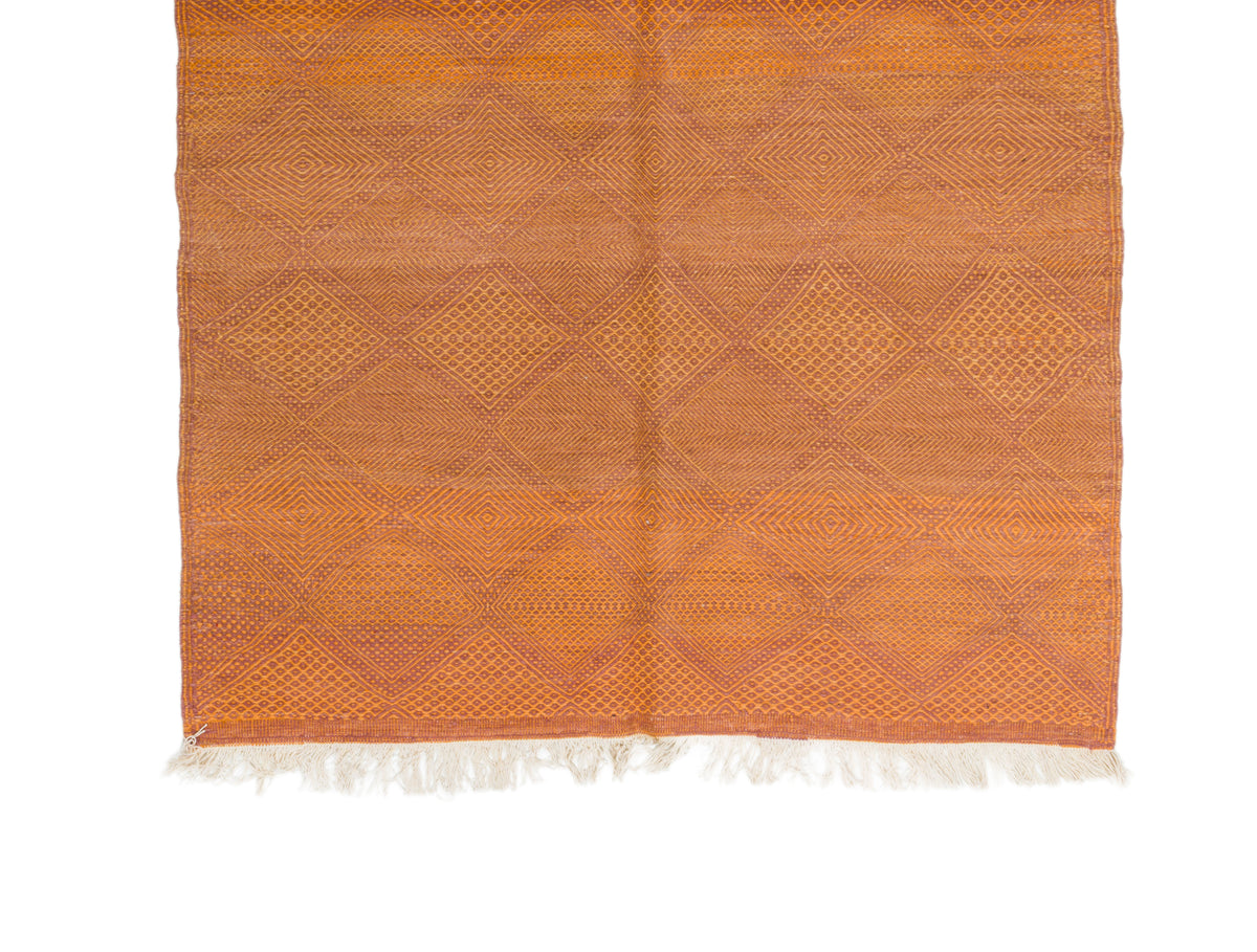 An orange woven area rug with a geometric diamond pattern and white fringe on the bottom edge, shown flat against a white background.