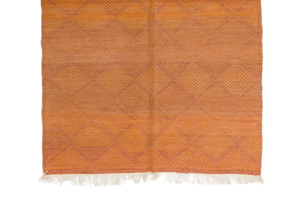 An orange woven area rug with a geometric diamond pattern and white fringe on the bottom edge, shown flat against a white background.