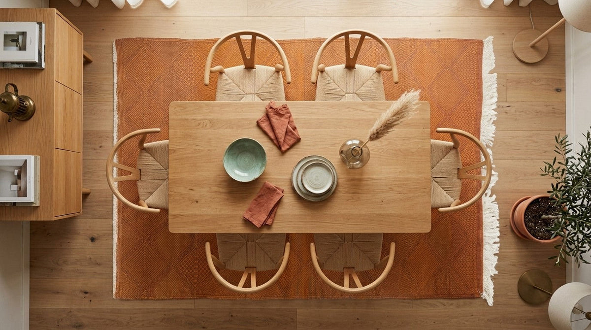 A top-down view of a wooden dining table surrounded by six woven-back chairs on a rust-orange Zanafi area rug, set in a warm, modern dining room with light wood floors.