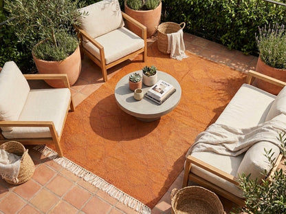 An outdoor patio seating arrangement featuring a large terracotta orange woven area rug under a round stone coffee table, surrounded by light wood-framed armchairs and a daybed with off-white cushions and a draped throw blanket.