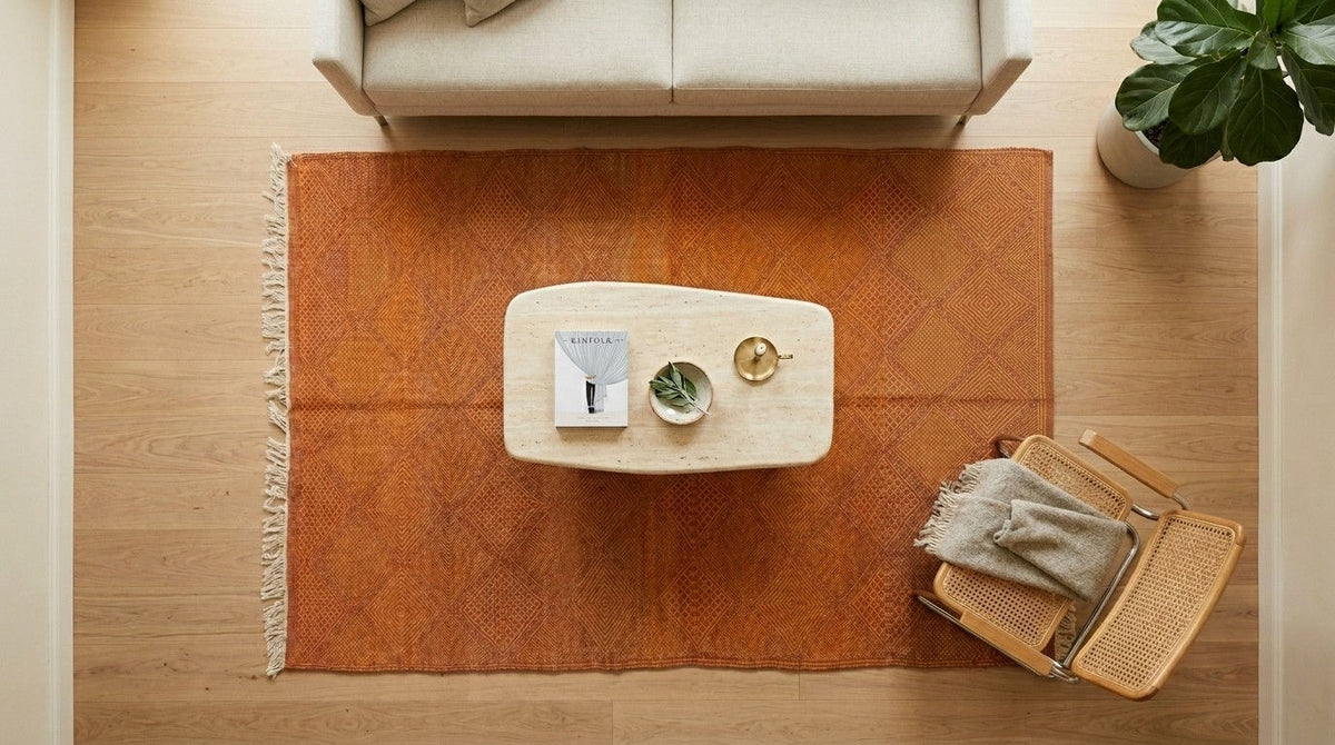 An overhead view of a warm rust-orange area rug styled in a modern living room setting with a light wood floor, a cream sofa, a small white coffee table, and a woven chair.