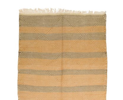 A rectangular wovenrug with fringe on one end, featuring horizontal stripes and a diamond lattice pattern in warm tan, beige, and taupe tones on a white background.