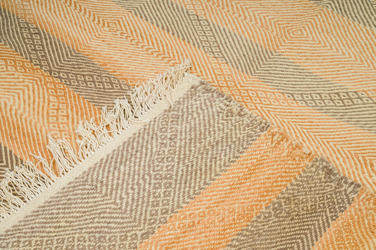 A close-up of a woven rug with a geometric pattern in peach, beige, and gray, featuring a fringed edge.