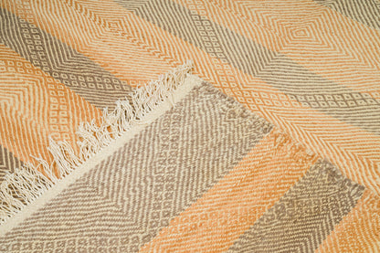 A close-up of a woven rug with a geometric pattern in peach, beige, and gray, featuring a fringed edge.