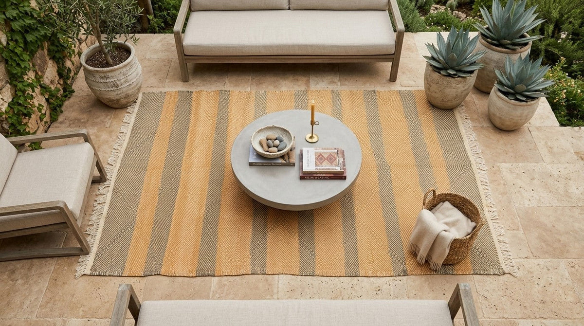 A superb Moroccan Zanafi rug set within a styled outdoor patio scene featuring a large rectangular area rug with vertical mustard yellow and taupe stripes, styled with a round coffee table, outdoor seating, and potted succulents.