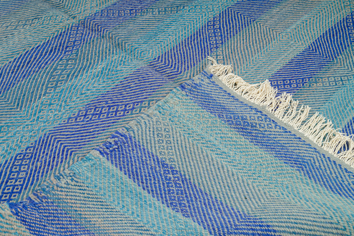 A close-up of a Moroccan Zanafi rug with a geometric pattern in shades of blue, light blue, and white, featuring white fringe on one end.