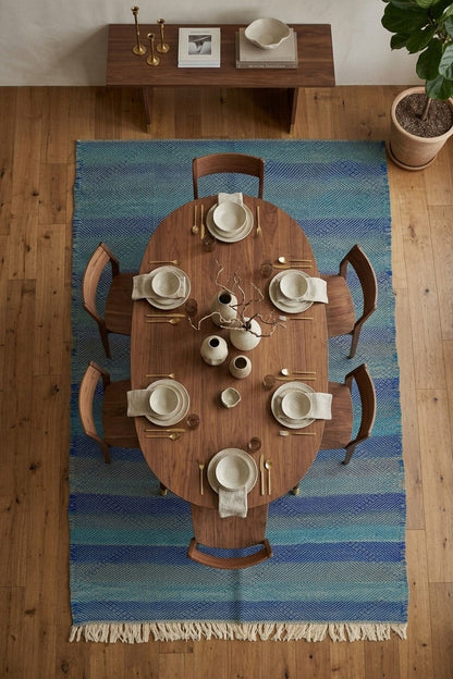 An overhead view of a dining room setup featuring a round wooden table set with white ceramic dishes and napkins, surrounded by six wooden chairs on a blue and teal striped woven area rug with cream fringe, placed on a light wood floor.