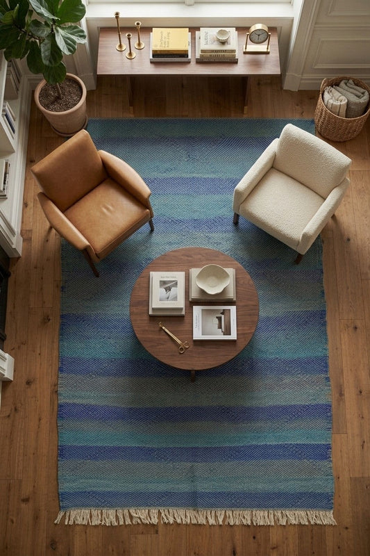 A top-down view of a blue and teal striped woven area rug with fringe, styled in a cozy living room with a round wooden coffee table, a brown leather armchair, and a cream armchair on a hardwood floor.