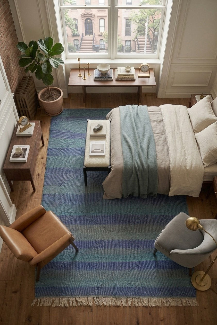 A top-down view of a styled bedroom featuring a prominent striped area rug in shades of blue, teal, and navy with cream fringe. The room includes a bed with neutral bedding, a leather armchair, a gray accent chair, and a large window looking out onto city buildings.