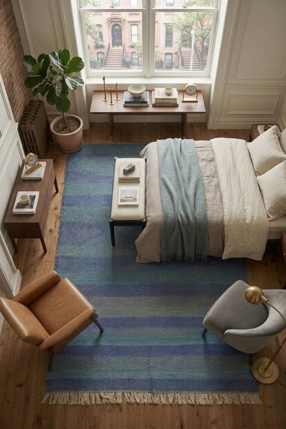 A top-down view of a styled bedroom featuring a prominent striped area rug in shades of blue, teal, and navy with cream fringe. The room includes a bed with neutral bedding, a leather armchair, a gray accent chair, and a large window looking out onto city buildings.