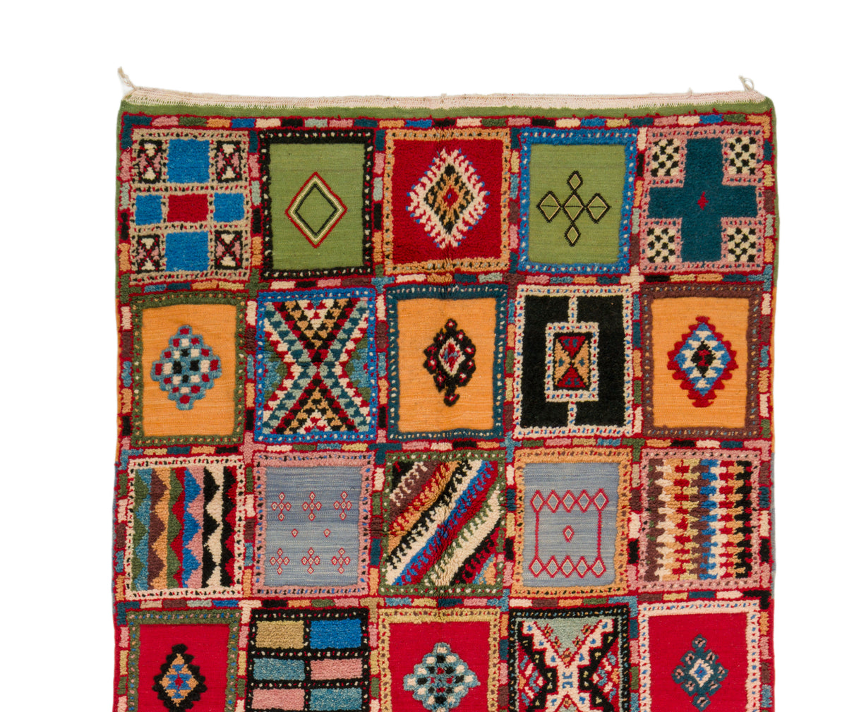 A handwoven rug featuring a patchwork design of colorful geometric squares with diamond, cross, and striped patterns, isolated on a white background.