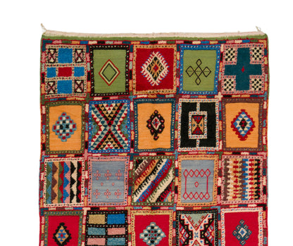 A handwoven rug featuring a patchwork design of colorful geometric squares with diamond, cross, and striped patterns, isolated on a white background.