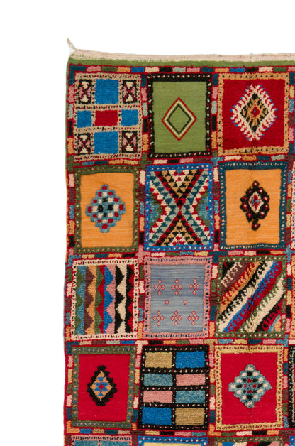 A close-up of a colorful patchwork-style woven rug or textile with multiple square panels in red, blue, green, yellow, orange, and black geometric patterns on a white background.