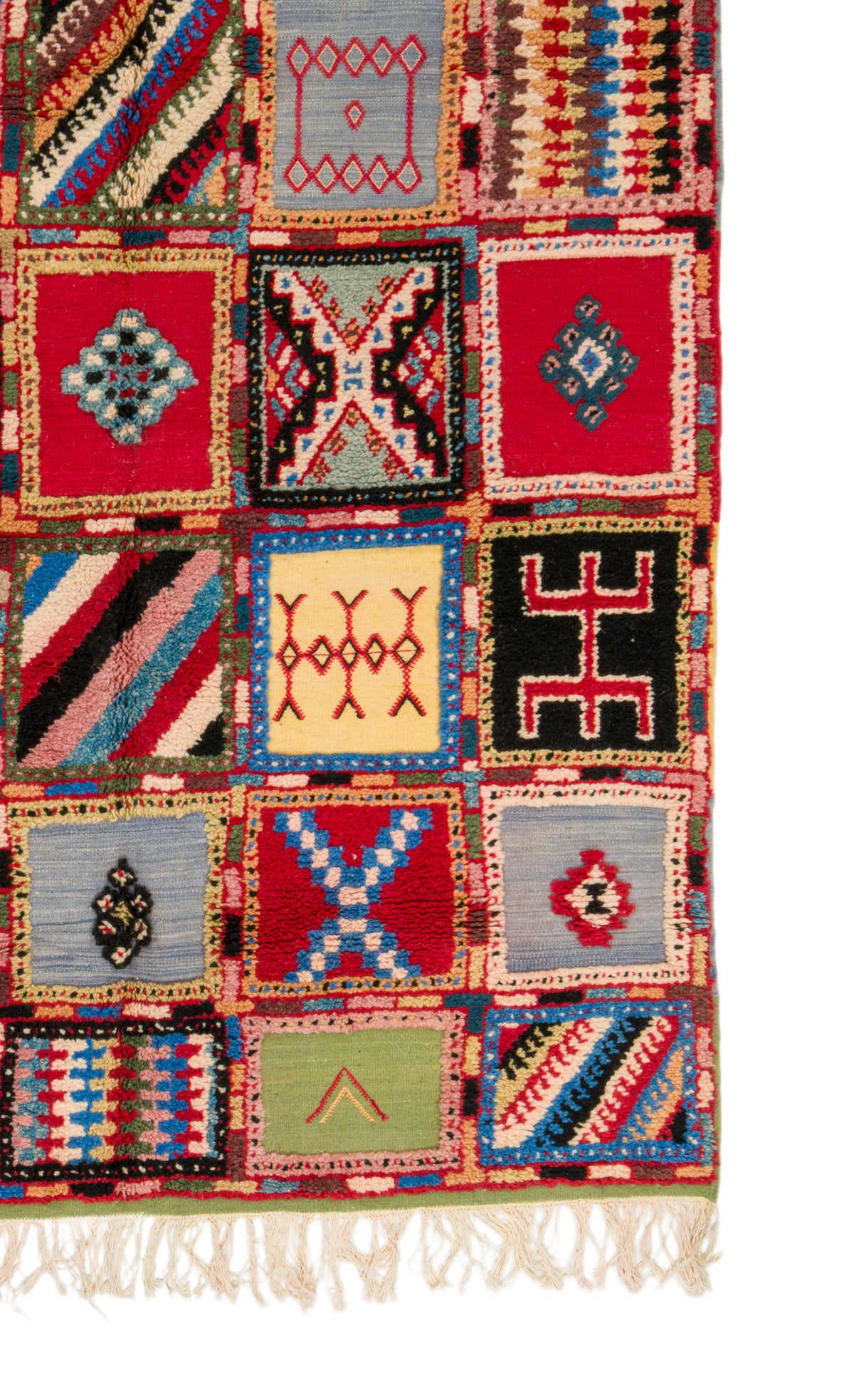 A close-up of a colorful patchwork-style woven rug with geometric and tribal-inspired square panels in red, blue, black, beige, green, and cream, finished with fringe along the bottom edge.