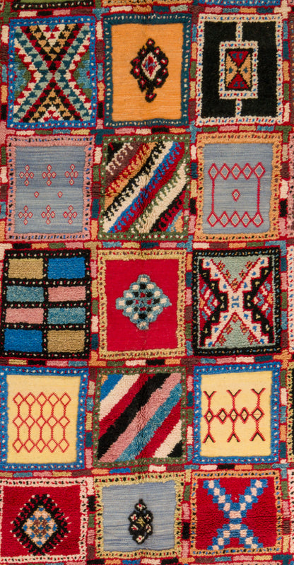 A close-up of a colorful patchwork rug featuring multiple square panels with geometric and tribal-inspired patterns in red, blue, yellow, black, beige, and pink.