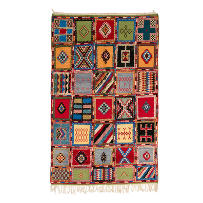 A multicolor patchwork-style woven rug with geometric diamond and striped motifs in red, blue, green, black, beige, and orange, finished with white fringe on the top and bottom, shown against a plain white background.