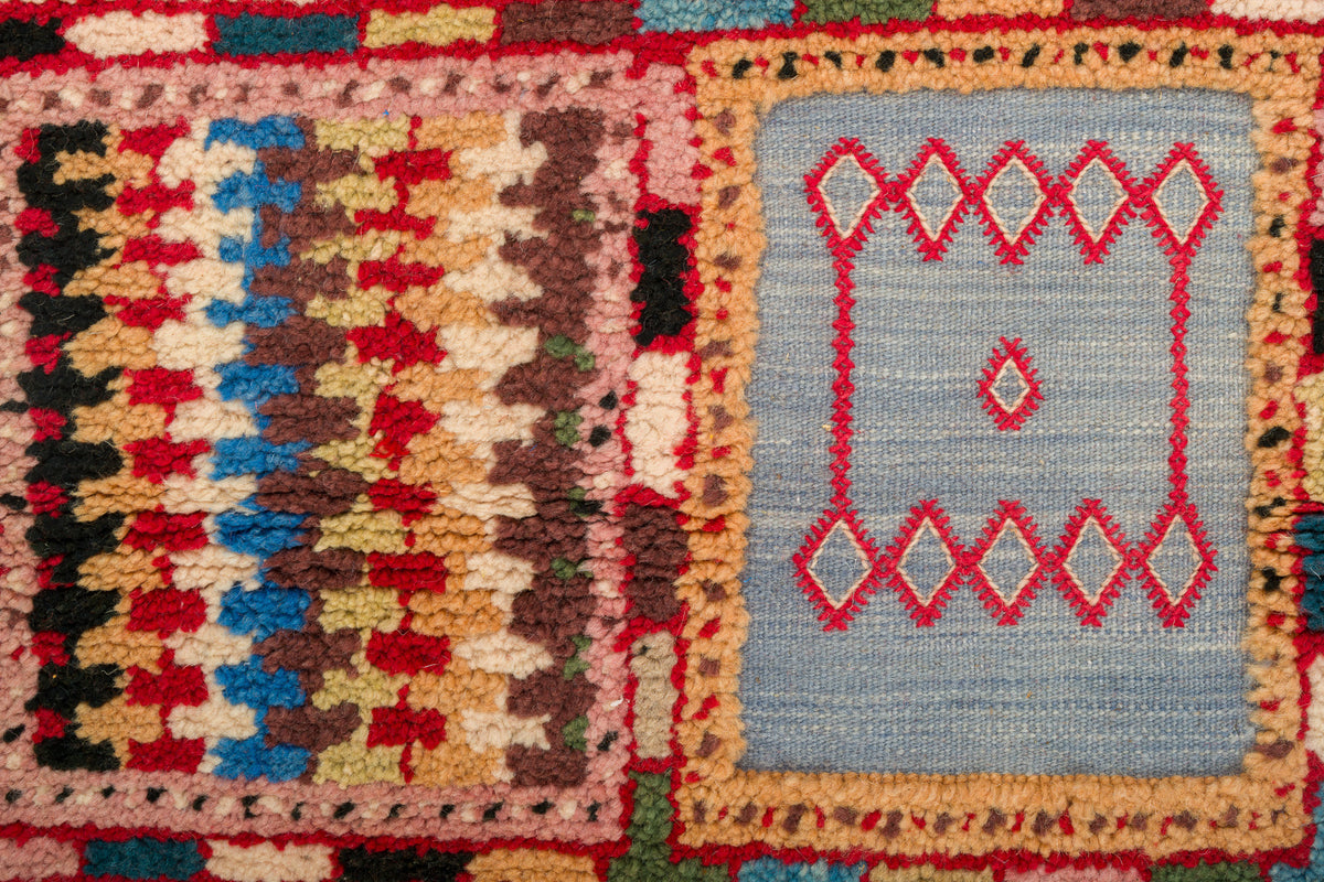 Close-up of a woven rug with two geometric panels: a left panel in red, beige, brown, black, and blue, and a right panel in light blue with red diamond motifs.