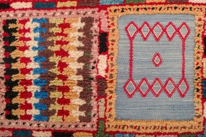 Close-up of a woven rug with two geometric panels: a left panel in red, beige, brown, black, and blue, and a right panel in light blue with red diamond motifs.