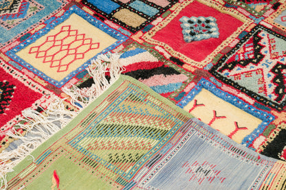 A close-up of a colorful woven rug with patchwork-style square panels, fringe on one side, and multiple hues including red, blue, beige, green, black, and white.