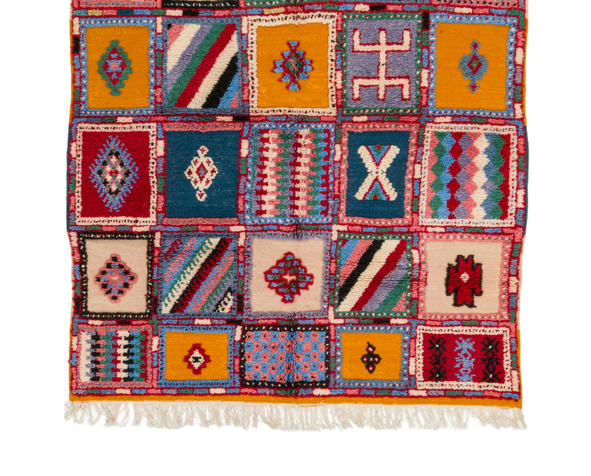 A Moroccan woven rug with a patchwork design of colorful geometric and tribal-inspired squares, finished with white fringe on the bottom edge.