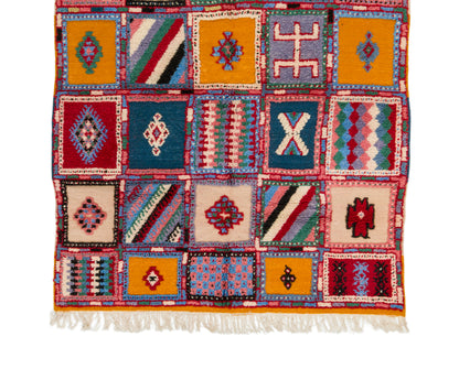 A Moroccan woven rug with a patchwork design of colorful geometric and tribal-inspired squares, finished with white fringe on the bottom edge.