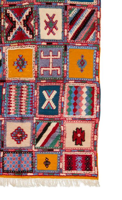 A close-up of a colorful patchwork-style woven rug with geometric and tribal-inspired square panels in red, blue, yellow, cream, and pink, finished with white fringe along the bottom edge.