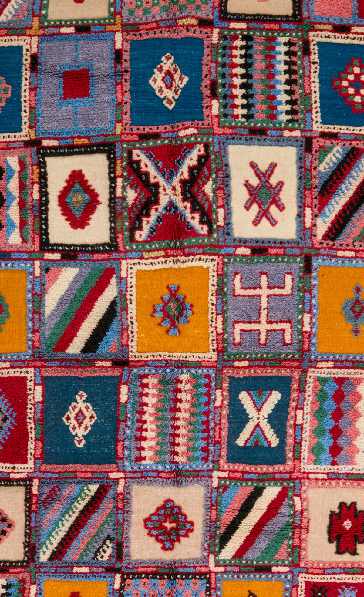 A close-up of a colorful geometric patchwork textile or rug featuring multiple square panels in blue, red, yellow, cream, black, and white with woven tribal-style patterns.