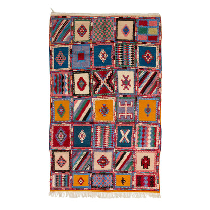 A rectangular handwoven kilim rug with a patchwork design of colorful geometric squares and a white fringed edge, shown flat against a white background.