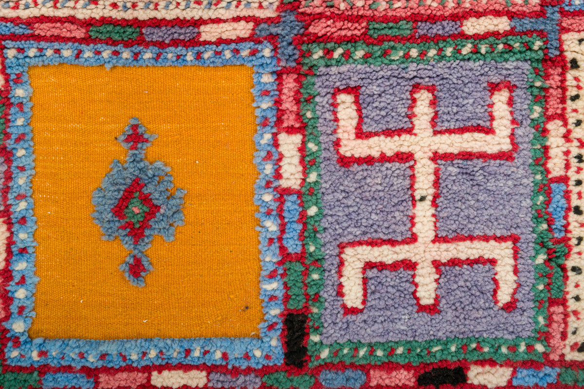 Close-up of a colorful woven rug with geometric panels, including a mustard yellow square with a blue diamond motif and a lavender square with a red geometric symbol.