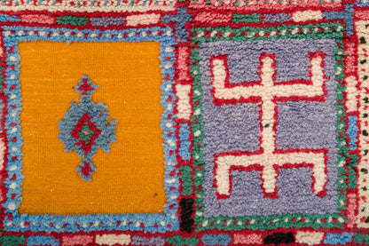 Close-up of a colorful woven rug with geometric panels, including a mustard yellow square with a blue diamond motif and a lavender square with a red geometric symbol.