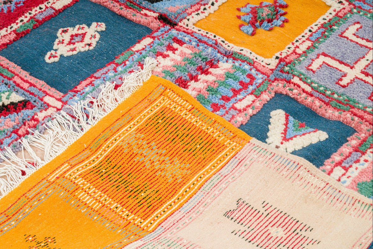 A close-up of a colorful patchwork-style woven rug with geometric and floral motifs in blue, orange, pink, cream, and red, including white fringe on one side.
