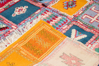 A close-up of a colorful patchwork-style woven rug with geometric and floral motifs in blue, orange, pink, cream, and red, including white fringe on one side.
