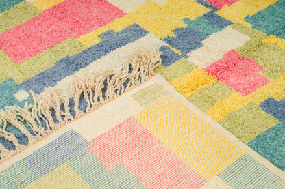 A close-up of a colorful geometric area rug with cream fringe, featuring blocks of pink, yellow, blue, green, and cream.