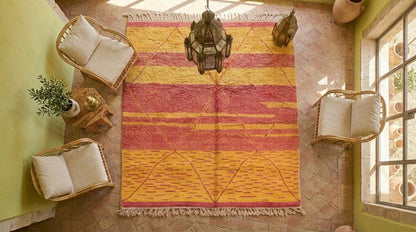 A large Moroccan Beni Ourain rug with a geometric pattern in orange, yellow, and pink, styled in a bright room with wicker chairs, a hanging lantern, and a tiled floor.