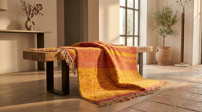 A colourful Beni Ourain Rug from Morocco with fringe, draped over a rustic wooden bench in a bright, modern interior room with warm sunlight and tiled floors.