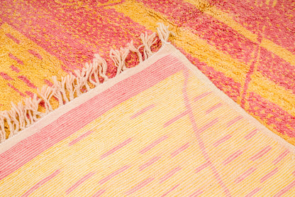 A close-up of a Beni Ourain Moroccan rug with cream fringe, showing a patterned surface in pink, yellow, and orange tones.