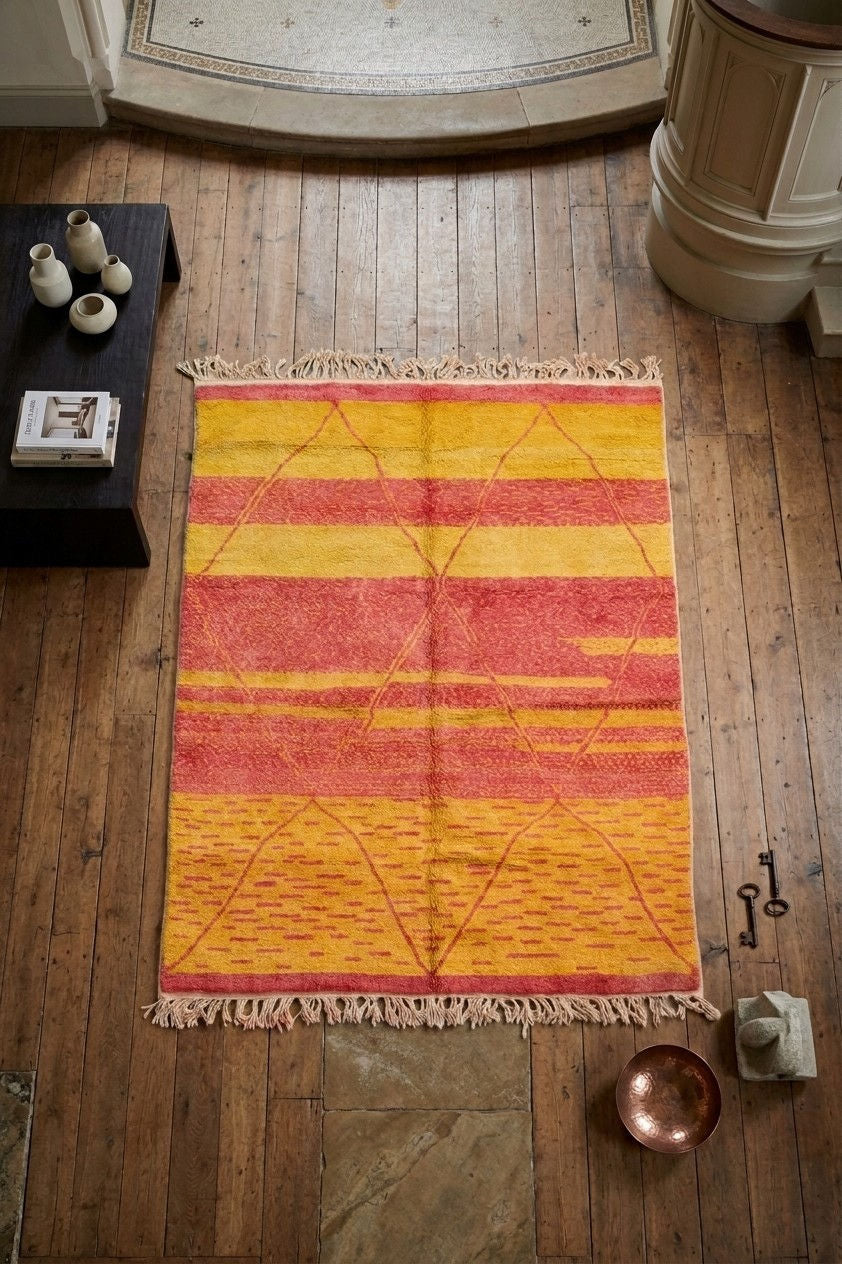A Beni Ourain rug with fringe, featuring horizontal stripes in yellow, orange, and red with a geometric diamond pattern, displayed on a rustic wooden floor.