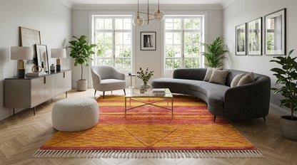 A modern living room featuring a prominent orange and red patterned Beni Ourain rug from Morocco, styled with a dark gray curved sectional sofa, a light gray armchair, and a sideboard.