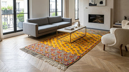 A yellow and orange patterned area rug with fringe, styled in a modern living room with a gray sofa, white armchair, and wood herringbone floor.