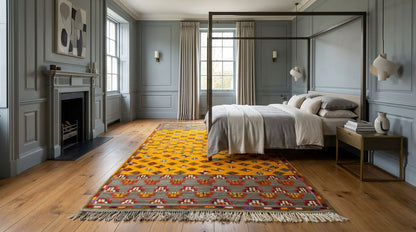 A large woven area rug with a yellow and orange geometric pattern and a multicolor fringe, styled on a wooden floor in a modern bedroom with a canopy bed and blue-gray paneled walls.