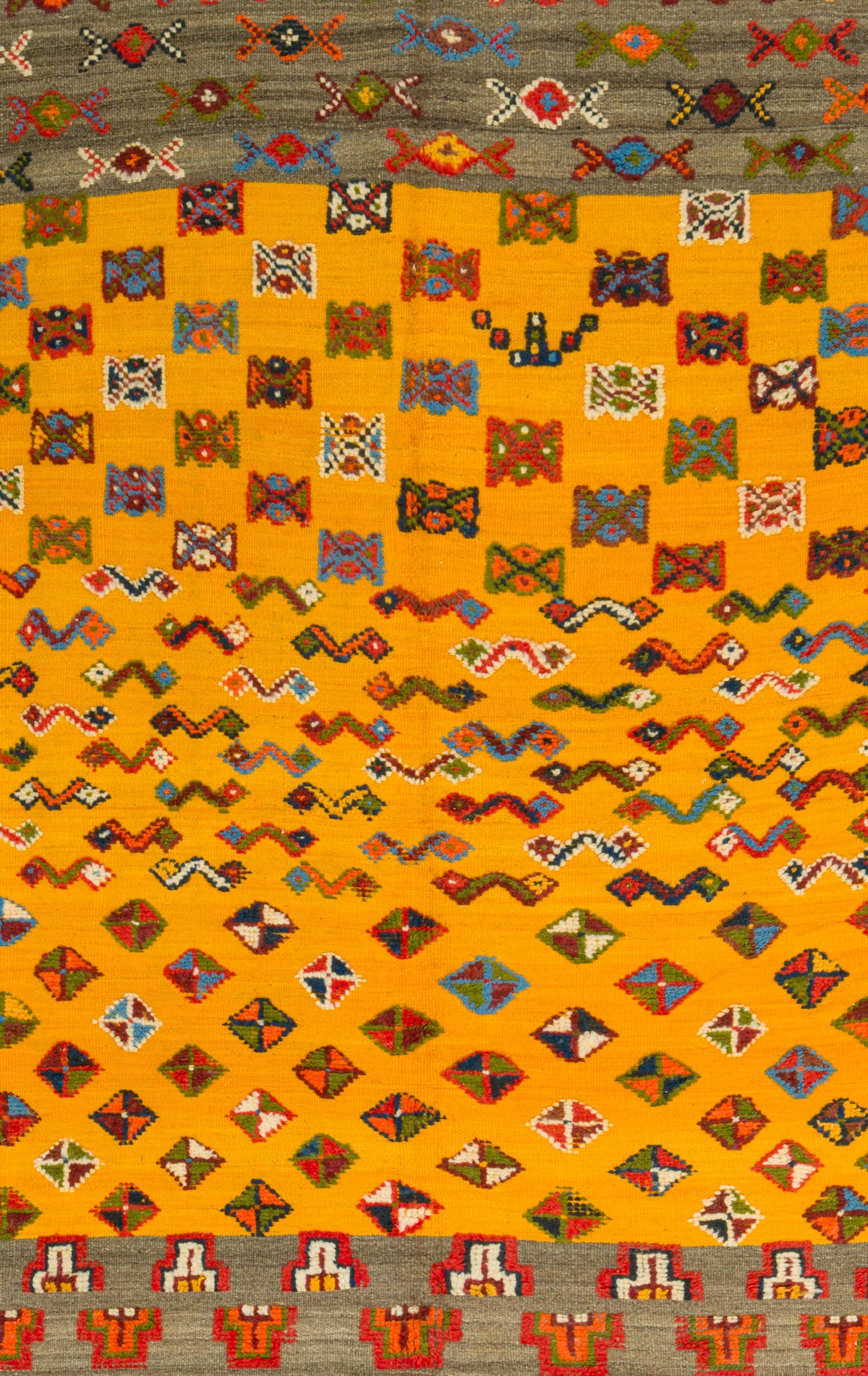 A close-up of a woven textile or rug featuring a bright yellow-orange field filled with rows of multicolored geometric and zigzag motifs, bordered by gray bands with red, white, and blue decorative patterns.